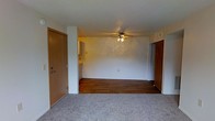 This single-floor residential space offers a straightforward and functional layout designed for comfortable everyday living. The main living area greets you upon entry through a warm wood-toned front door, immediately revealing an open and airy room that combines carpeted and hardwood flooring — a practical blend that defines distinct zones within the space. A ceiling fan with integrated lighting is centrally mounted overhead, providing both air circulation and ambient illumination throughout the area. The neutral, light-colored walls lend the space a clean and versatile backdrop, making it easy to personalize with furnishings and décor. A glimpse into the adjacent kitchen is visible from the living area, suggesting a connected and accessible layout that supports easy movement between spaces. An additional doorway on the far wall leads to further rooms within the unit, hinting at a well-organized floor plan. The overall ambiance is simple and move-in ready, with a calm, understated character well-suited for those seeking a no-frills, functional living environment. The combination of hard and soft flooring surfaces adds a degree of warmth and practicality to the space as a whole.