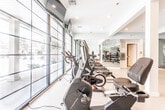 Located at 38 Old Short Hills Rd in Livingston, New Jersey, this single-level fitness center offers a well-appointed workout environment designed to support a broad range of exercise routines. The space is organized into distinct functional zones, allowing for both cardio training and strength work without crowding.

The cardio area is lined with treadmills, elliptical machines, and stationary bikes positioned along a wall of large, grid-pane windows that flood the floor with natural light and provide views of the surrounding exterior. Mounted flat-screen televisions above the equipment offer added convenience during workouts. Light hardwood-style flooring runs throughout, contributing to a clean, modern aesthetic complemented by soft mint-green walls and recessed ceiling lighting.

A dedicated strength and free weights section features a full dumbbell rack, medicine balls, resistance bands, stability balls, and a cable machine, all arranged alongside floor-to-ceiling mirrors that visually expand the space. A separate room, labeled Studio B, opens off the main floor through a glass-panel door and provides a more private setting for focused training or group sessions. Built-in shelving and a hydration station add practical amenities throughout the facility.