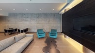 The Pearl's single-level amenity space showcases a sophisticated industrial-modern aesthetic throughout its open-plan layout. The entrance welcomes visitors with soaring ceilings, exposed ductwork, and expansive floor-to-ceiling windows that flood the interior with natural light while offering views of an outdoor courtyard. Large-format tile flooring runs seamlessly throughout, unifying the various zones within the space.

The lounge area features a striking design contrast between a large concrete-textured accent wall and a bold dark horizontal-paneled feature wall, accented by warm cylindrical uplights. Plush teal velvet armchairs and a grey sofa create inviting conversation clusters, complemented by sculptural accent tables. A billiards table anchors a dedicated recreation zone, while additional seating arrangements accommodate a variety of social configurations.

The fully equipped kitchen is defined by sleek dark cabinetry, a mosaic tile backsplash, and bright white countertops. A generous island with bar-height white modern stools serves as both a functional workspace and a casual gathering point. Pendant lighting illuminates the island, while recessed lighting enhances the overall brightness of the kitchen zone. The cohesive design palette of dark, white, and concrete tones delivers a refined yet functional communal environment.