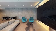 The Pearl's single-level amenity space showcases a sophisticated industrial-modern aesthetic throughout its open-plan layout. The entrance welcomes visitors with soaring ceilings, exposed ductwork, and expansive floor-to-ceiling windows that flood the interior with natural light while offering views of an outdoor courtyard. Large-format tile flooring runs seamlessly throughout, unifying the various zones within the space.

The lounge area features a striking design contrast between a large concrete-textured accent wall and a bold dark horizontal-paneled feature wall, accented by warm cylindrical uplights. Plush teal velvet armchairs and a grey sofa create inviting conversation clusters, complemented by sculptural accent tables. A billiards table anchors a dedicated recreation zone, while additional seating arrangements accommodate a variety of social configurations.

The fully equipped kitchen is defined by sleek dark cabinetry, a mosaic tile backsplash, and bright white countertops. A generous island with bar-height white modern stools serves as both a functional workspace and a casual gathering point. Pendant lighting illuminates the island, while recessed lighting enhances the overall brightness of the kitchen zone. The cohesive design palette of dark, white, and concrete tones delivers a refined yet functional communal environment.
