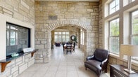 Located at 546 Frontier Rd in Lawrence, Kansas, this single-story club house is defined by its striking natural stone construction, which carries seamlessly from the exterior through every interior space. The architectural centerpiece is a grand stone archway connecting the main lounge to the dining and gathering areas, lending a timeless, castle-like character to the interior.

The lounge area features soaring ceilings, large multi-pane windows that flood the space with natural light, and a handsome stone fireplace topped with a flat-screen television — creating an inviting setting for relaxation and social gatherings. Adjacent to the lounge, a dedicated dining area accommodates a large farmhouse-style table and chairs beneath equally impressive high ceilings and expansive windows with outdoor views.

A kitchenette area serves residents with granite countertops, a bar-height seating counter, and a stone archway accent, offering a convenient refreshment station. A doorway from the kitchenette leads directly into the fitness center, which is a generously sized, well-lit space with soaring ceilings, clerestory windows, and rubberized flooring. The gym is outfitted with a comprehensive selection of strength and cardio equipment, including cable machines, weight racks, treadmills, and ellipticals, supported by structural stone columns that echo the building's distinctive design throughout.