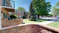 This single-floor property is situated within a thoughtfully landscaped residential community that exudes a welcoming, established character. The exterior is clad in classic red brick, lending the building a timeless and durable aesthetic. Curved brick garden walls in the foreground add architectural interest and define the outdoor spaces, hinting at attention to detail throughout the grounds.

The surrounding landscape is lush and well-maintained, featuring a mix of mature deciduous and evergreen trees that provide shade and visual depth. A neatly trimmed lawn stretches across the communal grounds, contributing to an open, park-like atmosphere. Additional units visible in the background suggest this property is part of a larger residential community, with balconies and large windows offering residents both outdoor access and natural light.

The paved pathways and parking areas are neatly integrated into the landscape, ensuring practical accessibility while preserving the green surroundings. The overall ambiance is calm and residential, combining classic brick architecture with mature, verdant landscaping to create an inviting and well-established environment.