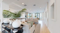 Located at 1464 Laurel Sound Ln in Sanford, FL, this expansive single-floor space offers a thoughtfully designed open-concept layout that seamlessly integrates a dining room, kitchen, and living room into one cohesive environment. The generously proportioned floor plan provides an airy, flowing sense of space throughout.

The living area is anchored by neutral linen sofas, distinctive live-edge wood coffee tables, and lush tropical plants that reinforce a relaxed coastal aesthetic. Recessed lighting and large floor-to-ceiling windows flood the interiors with abundant natural light, while light-toned hardwood flooring runs continuously across the space, enhancing the sense of openness.

A dramatic architectural highlight is the grand lattice-style wood beam ceiling structure in the central zone, creating visual depth and defining the dining area below, which features rich dark wood tables paired with warm leather seating. A standout lounge nook features suspended swing-style seating and a vivid tropical mural, adding a playful, resort-inspired focal point.

The hallway connects the various functional zones and leads to a striking staircase with light wood treads and sleek black metal railings set against soaring double-height windows. Throughout, crisp white walls, natural wood accents, and teal-framed exterior doors deliver a modern yet inviting atmosphere with a distinctly Florida-inspired character.