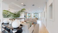 Located at 1464 Laurel Sound Ln in Sanford, FL, this expansive single-floor space offers a thoughtfully designed open-concept layout that seamlessly integrates a dining room, kitchen, and living room into one cohesive environment. The generously proportioned floor plan provides an airy, flowing sense of space throughout.

The living area is anchored by neutral linen sofas, distinctive live-edge wood coffee tables, and lush tropical plants that reinforce a relaxed coastal aesthetic. Recessed lighting and large floor-to-ceiling windows flood the interiors with abundant natural light, while light-toned hardwood flooring runs continuously across the space, enhancing the sense of openness.

A dramatic architectural highlight is the grand lattice-style wood beam ceiling structure in the central zone, creating visual depth and defining the dining area below, which features rich dark wood tables paired with warm leather seating. A standout lounge nook features suspended swing-style seating and a vivid tropical mural, adding a playful, resort-inspired focal point.

The hallway connects the various functional zones and leads to a striking staircase with light wood treads and sleek black metal railings set against soaring double-height windows. Throughout, crisp white walls, natural wood accents, and teal-framed exterior doors deliver a modern yet inviting atmosphere with a distinctly Florida-inspired character.
