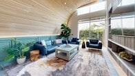 Located at 27930 Cabot Rd in Laguna Niguel, CA, this thoughtfully designed single-floor space offers a generously sized, open-concept layout that blends modern architecture with warm, inviting finishes. The interior is immediately distinguished by a dramatic arched slatted wood ceiling that adds organic texture and curves gracefully over a sophisticated lounge area. Wide-plank light wood flooring runs throughout, complementing the bright, airy atmosphere created by floor-to-ceiling windows and sheer drapery that flood the space with natural light.

A defining architectural feature is the bold steel-framed glass partition wall, which elegantly separates the lounge seating area from the open communal space while maintaining visual connectivity throughout. The lounge showcases teal wainscoting, plush blue leather seating, and an artisan area rug, creating a refined yet comfortable gathering environment. A separate dining area provides a round glass-top table with contemporary blue-and-gold chairs, set against expansive windows overlooking lush, manicured greenery and palm trees.

The open layout also accommodates a game area, visible from the main space, reinforcing the multi-functional nature of this community amenity. Abundant greenery, including potted fiddle-leaf fig and tropical plants, enhances the fresh, California-inspired ambiance throughout.
