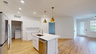 Located at 7015 Market St in Wilmington, NC, this thoughtfully designed 2-bedroom, 3-bathroom residence offers a clean, modern aesthetic throughout its single-floor layout. The open-concept living and kitchen area serves as the heart of the home, anchored by a central island with a stainless steel sink, dishwasher, and stylish gold pendant lighting overhead. The kitchen is outfitted with crisp white shaker cabinets accented by gold hardware, a white subway tile backsplash, and a full suite of stainless steel appliances including a refrigerator, range, and microwave. Light hardwood flooring flows throughout the main living areas, enhancing the bright, airy atmosphere created by recessed lighting and white walls. The spacious primary bedroom features soft carpeting, a ceiling fan, and a walk-in closet with built-in wire shelving for ample storage. Additional bedrooms are equally well-sized with carpeted floors and natural light. Three full bathrooms provide convenience, including a tub/shower combo. A dedicated laundry room with full-size washer and dryer adds everyday practicality. With three closets total and a clean, contemporary design, this residence delivers functional comfort in a polished Wilmington setting.