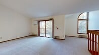 Located at 3600 S Rivershire Drive in Greenfield, Wisconsin, this single-floor unit offers a well-organized layout designed for comfortable living. The expansive living room is carpeted and bathed in natural light, featuring a wood-trimmed sliding glass door that opens to a private balcony overlooking mature trees — a serene outdoor retreat. A staircase with wooden railings connects the level, and an additional window provides supplemental natural light to the living space. The kitchen is efficiently designed with warm oak cabinetry, light countertops, tile flooring, and a full suite of black appliances including a refrigerator, electric range, microwave, and dishwasher. A pass-through counter connects the kitchen to the adjacent living area, enhancing the open feel. The bedrooms are carpeted and neutral-toned, with one room featuring sliding wood-paneled closet doors offering generous storage capacity, and another with a wood-trimmed window and baseboard heating. The bathroom includes tile flooring, a wood-vanity with single-sink, a mirror with overhead lighting, and clean, functional fixtures. Throughout the unit, warm wood trim accents complement the light wall tones, creating a cohesive and inviting atmosphere.