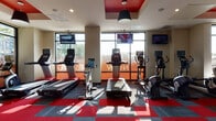 Located at 3833 S Bond Ave in Portland, Oregon, this well-appointed fitness center delivers a dynamic and energetic workout environment designed to meet a wide range of exercise needs. The space is defined by its bold aesthetic, featuring a striking red-and-gray modular carpet floor pattern that complements the vivid red accent panels integrated into the ceiling design. Bold pendant lights hang from the ceiling alongside recessed lighting, creating a bright and motivating atmosphere throughout.

Large floor-to-ceiling windows line multiple walls of the facility, flooding the space with natural light and offering open views of the surrounding outdoor landscape. The cardio zone is generously equipped with treadmills, upright and recumbent bikes, elliptical trainers, and rowing machines, each outfitted with individual digital displays. Wall-mounted flat-screen televisions are positioned throughout for added convenience during workouts.

A dedicated free weights area features a full rack of dumbbells in a range of sizes, accompanied by weight benches for strength training. A multi-functional cable machine system provides options for a comprehensive resistance workout. An additional enclosed section, separated by glass-paneled partitions, houses specialty equipment including medicine balls, resistance tools, and other functional fitness accessories. The combination of abundant natural light, modern equipment, and thoughtful design makes this fitness center a well-rounded facility.