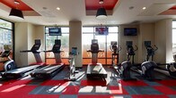 Located at 3833 S Bond Ave in Portland, Oregon, this well-appointed fitness center delivers a dynamic and energetic workout environment designed to meet a wide range of exercise needs. The space is defined by its bold aesthetic, featuring a striking red-and-gray modular carpet floor pattern that complements the vivid red accent panels integrated into the ceiling design. Bold pendant lights hang from the ceiling alongside recessed lighting, creating a bright and motivating atmosphere throughout.

Large floor-to-ceiling windows line multiple walls of the facility, flooding the space with natural light and offering open views of the surrounding outdoor landscape. The cardio zone is generously equipped with treadmills, upright and recumbent bikes, elliptical trainers, and rowing machines, each outfitted with individual digital displays. Wall-mounted flat-screen televisions are positioned throughout for added convenience during workouts.

A dedicated free weights area features a full rack of dumbbells in a range of sizes, accompanied by weight benches for strength training. A multi-functional cable machine system provides options for a comprehensive resistance workout. An additional enclosed section, separated by glass-paneled partitions, houses specialty equipment including medicine balls, resistance tools, and other functional fitness accessories. The combination of abundant natural light, modern equipment, and thoughtful design makes this fitness center a well-rounded facility.