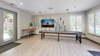 Located at 3301 Arena Blvd in Sacramento, CA, this thoughtfully designed single-floor suite offers a cohesive blend of comfort and functionality across three distinct spaces. The hallway serves as a stylish welcoming area, featuring light wood-look flooring, neutral-toned walls accented by a bold dark green geometric detail, framed botanical artwork, and cozy lounge seating arranged symmetrically on either side — creating an inviting and polished first impression. Beyond the hallway, a spacious recreation and lounge area stands out with its eye-catching accent wall of multicolored vertical panels flanking a large wall-mounted flat-screen TV. A full-size shuffleboard table and a high bar-height table with stools make this an ideal gathering space, complemented by natural light filtering through shuttered windows. The private office is equipped with built-in wraparound desks, ergonomic seating, and plantation shutters that allow ample natural light, making it a productive workspace. Completing the suite, the kitchenette features white cabinetry, dark countertops, a stainless steel sink, and a full-size stainless steel refrigerator — providing convenient amenities for everyday use. Throughout the space, light wood flooring and a neutral color palette tie each area together seamlessly.