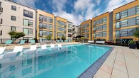This outdoor amenity space is a standout feature of a modern residential community, defined by its striking contemporary architecture. The building exterior showcases a bold combination of warm yellow and cool grey siding panels, floor-to-ceiling windows, and private balconies spanning multiple stories — creating an enveloping courtyard-like atmosphere around the amenity deck.

At the heart of the space is a generously sized resort-style swimming pool with crystal-clear blue water, surrounded by a spacious tiled pool deck finished in warm neutral tones. The deck is thoughtfully furnished with an array of white in-pool lounge chairs, teal and charcoal chaise lounges, and comfortable upholstered outdoor seating with side tables — ideal for relaxing in the open air. Shade options include a large patio umbrella positioned near a seating cluster at the pool's edge.

Adjacent to the main pool, a separate spa offers an inviting hot tub with easy step-in access, accompanied by additional lounge seating. Tall black rectangular planters with mature trees add greenery and structure to the deck, while iron perimeter fencing provides definition without obstructing the open-sky views. The overall ambiance is modern, resort-inspired, and well-appointed, offering a refined outdoor retreat within the community.