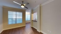 This single-floor residence showcases a thoughtfully designed open-concept layout that seamlessly connects the living area to the kitchen. The living space is finished with rich hardwood-style flooring that runs throughout, complementing the warm neutral tones of the walls. Crown molding along the ceiling adds a refined architectural detail, while a ceiling fan with a built-in light fixture provides both functionality and a classic aesthetic.

Natural light is a defining feature of this space, with multiple windows fitted with horizontal blinds allowing for adjustable brightness and privacy. The arched pass-through opening between the living area and kitchen serves as a stylish architectural element, framing a dark-toned countertop bar that offers a casual dining or workspace option. Track lighting above the kitchen area ensures ample task illumination.

The kitchen, visible through the arched opening, includes a sink and continues the cohesive design language of the rest of the unit. Clean white baseboards and trim throughout provide a crisp contrast to the warm flooring and wall tones, contributing to an overall ambiance that feels polished, bright, and move-in ready.