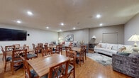 This single-floor community space is thoughtfully designed to serve as a versatile gathering hub, seamlessly blending a fully equipped kitchen, a generous dining area, and a relaxed lounge into one open-concept layout. The kitchen is a standout feature, boasting white cabinetry, a white subway tile backsplash, stainless steel appliances including a range, dishwasher, and refrigerator, and a large central island topped with striking granite countertops. Pendant lights hang above the island, adding warmth and definition to the cooking and prep area. The expansive dining section accommodates multiple wooden tables and cushioned chairs arranged to encourage group meals and gatherings, all bathed in recessed lighting overhead. A wall-mounted flat-screen television anchors one end of the dining area, making it suitable for presentations or communal viewing. Warm hardwood-style flooring flows throughout the entire space, contributing to a cohesive and inviting atmosphere. At the far end, a tastefully appointed lounge area features plush sofas and accent chairs arranged around a glass coffee table, complemented by decorative artwork, table lamps, and layered area rugs. The overall ambiance is clean, functional, and welcoming — ideal for community events, shared meals, and casual socializing.