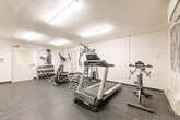 Located at 1363 Lakeshore Rd in Burlington, Ontario, this fitness room offers a well-organized and fully equipped workout space within a compact, single-level layout. The room features a rubber-tile floor designed to withstand heavy equipment use, white walls, and a textured ceiling fitted with fluorescent strip lighting that ensures consistent, bright illumination throughout the space. A wall-mounted split air conditioning unit provides climate control for year-round comfort during workouts.

The fitness area is outfitted with a variety of cardio equipment, including treadmills and an elliptical trainer, along with a multi-station cable machine that supports a wide range of strength exercises. A dedicated dumbbell rack with a full set of free weights, medicine balls, and a flat workout bench provide additional training versatility. An exercise step and stability ball further round out the available equipment.

A defining architectural feature of the room is the large panel of floor-to-ceiling mirrors mounted along one wall, which visually expand the space and allow users to monitor their form during exercise. Motivational black-and-white athletic photography is displayed on the walls, contributing to an energetic and focused ambiance. A floor fan is also on hand for additional ventilation. The room is accessed through a standard interior door, making it a convenient and self-contained fitness facility.