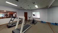 Located at 1515 Steffen Ct in Fort Wright, Kentucky, this single-floor fitness center is a well-equipped exercise facility designed to support a variety of cardio and general fitness activities. The space is laid out across a carpeted room with light gray walls, warm wood baseboards, and a textured ceiling fitted with recessed fluorescent lighting that keeps the area well-illuminated throughout. A large mirror spans one wall, visually expanding the space and allowing users to monitor their form during workouts. Natural light filters in through windows dressed with vertical blinds, adding a welcoming brightness to the room. The fitness area is furnished with multiple treadmills, an elliptical trainer, a stationary upright bike, and a step machine, offering a comprehensive range of cardio equipment. A wall-mounted television is positioned for easy viewing during exercise. Additional accessories, including exercise mats and resistance equipment, are stored along the walls for convenient access. A solid wood paneled door provides entry to the space, complementing the room's understated, functional aesthetic. A pass-through window connects to an adjacent room, suggesting access to additional amenities nearby. Overall, this fitness center offers a clean, practical, and comfortable environment suited for daily exercise.