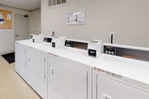This single-floor shared amenity space is thoughtfully designed to serve the practical and social needs of its users. The laundry area forms the heart of the facility, housing multiple commercial-grade washing machines and dryers arranged in efficient rows along the walls. The machines are complemented by wall-mounted informational panels and a bulletin board, keeping residents informed and organized. The flooring throughout the laundry area is a durable, easy-to-clean vinyl, well-suited for high-traffic use. Adequate overhead fluorescent lighting ensures a bright, functional environment. A laundry cart is also available for added convenience. Adjacent to the laundry area is a comfortable lounge and seating nook, featuring a long dark table with chairs, ideal for waiting or casual gatherings. A large window dressed with a decorative patterned valance fills the space with natural light and offers a pleasant view of the surrounding outdoor greenery. A Coca-Cola vending machine and a decorative potted plant add color and a welcoming touch to the space. The neutral wall tones and crown molding in the lounge area lend a clean, polished finish. Together, these areas create a practical yet inviting communal space.