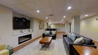 This single-floor space is designed as a welcoming communal lounge, combining functional amenities with a comfortable, contemporary aesthetic. The main seating area is anchored by a sleek, linear electric fireplace set into a white surround, paired with a wall-mounted flat-screen television above — creating a natural focal point for the room. Two dark leather sofas, accented with teal and neutral throw pillows, are arranged around a mid-century style wooden coffee table atop a patterned area rug, lending warmth and texture to the space.

The drop ceiling features a grid of recessed lighting that evenly illuminates the interior, compensating for the absence of large windows while maintaining a bright and inviting atmosphere. The flooring transitions from the carpeted lounge area to hardwood-style flooring toward the rear of the space, where a kitchenette or service counter is visible. This secondary area appears equipped for light food and beverage use, adding practical utility to the lounge. Neutral wall tones and a piece of abstract artwork contribute to the overall relaxed, modern ambiance. An emergency exit sign and corridor are visible at the rear, indicating this is a shared or commercial-use facility.