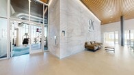 This contemporary facility is defined by thoughtful architectural design and high-quality finishes throughout. The grand entrance lobby sets the tone with soaring ceilings clad in warm wood slat panels, large-format light stone tile flooring, and a dramatic full-height feature wall adorned with an intricate tonal wave pattern and a prominent "M" emblem. Comfortable leather seating and floor-to-ceiling glass entry doors contribute to the welcoming, upscale atmosphere.

The open corridor extends from the lobby, flanked by the same signature feature wall and complemented by glass-panel railings with warm wood-capped handrails that overlook a lower lounge level furnished with seating and views of the surrounding landscape. Structural columns and elevator access points are integrated seamlessly into the design.

The expansive fitness center is a standout feature, offering a comprehensive array of Cybex cardio equipment — including treadmills, ellipticals, and bikes — alongside a robust strength-training section with cable machines and free weights. Vibrant geometric patterned carpet tiles in blue, teal, and warm earth tones energize the space, while floor-to-ceiling windows flood the gym with natural light and open views.

Dedicated studio rooms with light wood-look flooring, full-length mirrored walls, and wall-mounted displays provide flexible space for group fitness or personal training. The facility combines functionality with a polished, modern aesthetic throughout.
