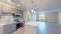 This well-appointed single-floor residence offers a thoughtfully designed open-concept layout that seamlessly connects the kitchen, dining, and living areas. The kitchen is a standout feature, showcasing gray shaker-style cabinetry that extends to the ceiling, a mosaic tile backsplash, and a full suite of stainless steel appliances including a refrigerator, electric range, built-in microwave, and dishwasher. A central island with a crisp white countertop provides additional prep space and serves as a natural gathering point, accented by stylish pendant lighting overhead. Crown molding runs throughout the space, adding a refined architectural detail to the neutral-toned walls. Light hardwood-style flooring flows continuously across the main living areas, enhancing the sense of openness and brightness. The spacious living room benefits from natural light through a large window and is finished with a ceiling fan. Arched doorways lend a classic touch to the transitional design. The bathrooms continue the clean, modern aesthetic with light flooring, dark vanity cabinetry, and white countertops. One bathroom includes safety grab bars, reflecting a practical and inclusive design. Track lighting in the kitchen and ceiling fixtures throughout ensure the space is well-lit at all hours.