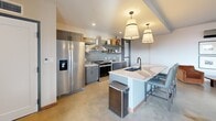 This thoughtfully designed single-floor space combines a modern kitchen, dining area, and living room in a seamless open-plan layout. The kitchen is anchored by a large central island topped with striking veined marble countertops, paired with gray cabinetry, a glossy subway tile backsplash, stainless steel appliances, and open shelving. Pendant lighting with cream drum shades provides warm, focused illumination throughout. Polished concrete floors run continuously across the entire interior, lending a sleek industrial character that ties each zone together cohesively. The living area features exposed wood ceiling beams, a dramatic slatted wood partition wall, and a fireplace set against a bold dark accent wall. A statement geometric chandelier hangs above the seating area, which benefits from floor-to-ceiling windows that flood the space with natural light and frame panoramic city views. The dining zone offers built-in banquette seating alongside bistro-style tables with richly veined marble tops, adding a lounge-like warmth. A generously sized outdoor terrace extends the living space further, furnished with ample seating and a dining table, all under a covered ceiling — ideal for taking in expansive views of the surrounding cityscape and green spaces below.