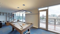 This single-floor entertainment space is designed for leisure and social gatherings, combining functional amenities with an elevated urban setting. At its center sits a rustic wood-framed pool table, positioned beneath a striking rectangular pendant light that frames the playing surface with clean, modern illumination. The blue carpeted flooring anchors the room and complements the contemporary aesthetic throughout.

Large floor-to-ceiling windows line the perimeter, flooding the space with abundant natural light while offering expansive views of the surrounding cityscape. A glass-paneled door provides direct access to an outdoor terrace, further connecting the interior to the urban panorama beyond. The lounge area, positioned near the windows, offers comfortable seating with views that enhance the relaxed atmosphere.

The neutral-toned walls and smooth ceiling maintain a clean, open feel, allowing the architectural elements and furnishings to take center stage. Recessed lighting supplements the pendant fixture, ensuring the space is well-lit throughout. Overall, this entertainment room delivers a polished, inviting environment that balances recreational use with sophisticated design and remarkable city views.