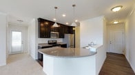 Located at 3381 Blue Heron Dr in Hilliard, Ohio, this single-story home offers a thoughtfully designed layout with modern finishes throughout. The kitchen serves as a central highlight of the space, showcasing rich dark cabinetry that provides ample storage and creates a striking contrast against the light granite countertops. A curved peninsula with a white base extends into the living area, offering additional prep space and a natural gathering spot. Stainless steel appliances, including a gas range, built-in microwave, and refrigerator, lend a sleek, contemporary feel to the kitchen. Pendant lighting hangs above the peninsula, complementing the recessed ceiling lights that illuminate the space. A built-in sink along the peninsula adds to the kitchen's functional design. The flooring transitions from dark hardwood in the kitchen to plush carpeting in the adjacent living areas, adding warmth and comfort. Crown molding details along the ceiling contribute an elegant, finished look throughout. The open layout promotes a seamless flow between the kitchen and living spaces, making this home well-suited for both everyday living and entertaining.