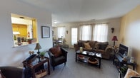 This single-floor residence offers a welcoming and functional layout designed for comfortable living. The living room serves as the heart of the home, featuring plush carpeted flooring, neutral-toned walls, and an abundance of natural light streaming through multiple windows dressed with sheer, elegant curtains. A glass-paneled door provides additional brightness and a connection to the outdoors. The living area is generously sized, accommodating a full sofa with decorative pillows, a leather armchair, and a glass-top coffee table, creating a cozy yet refined gathering space. A side console table with tasteful décor accents anchors one corner of the room. The space flows seamlessly into a dedicated dining area, visible through an open passageway, where a dining table and chairs are set beneath warm, ambient lighting. A pass-through window with warm lighting connects to an adjacent kitchen area, adding a charming architectural detail that enhances the open feel of the floor plan. Throughout, the design balances practicality with comfort, featuring consistent flooring, clean lines, and a warm, earthy color palette that gives the entire space a cohesive and inviting ambiance.