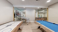 Located at 1881 E Irvington Rd in Tucson, AZ, this dedicated game room is a compact yet efficiently designed entertainment space. The room is finished with light hardwood-style flooring and crisp white walls, creating a clean, modern aesthetic that keeps the focus on the wide variety of games available.

The space is outfitted with an impressive selection of recreational equipment, including a pool table with a vibrant blue felt surface, an air hockey table with a natural wood base, a classic foosball table, and a skee-ball machine — offering entertainment options for guests of all ages and interests.

Recessed lighting throughout the ceiling ensures the room is well-lit for gameplay, while a wall-mounted mini-split air conditioning unit provides climate control for year-round comfort. A window along one wall allows for natural light and offers a view of the surrounding greenery outside.

Large wall mirrors are strategically placed and framed with natural wood accents, visually expanding the room and enhancing its open feel. A dedicated entrance door provides easy access to the space. The combination of thoughtful design, ample recreational equipment, and comfortable amenities makes this game room a highly functional and engaging destination for leisure and social activity.