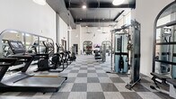 This well-appointed fitness center offers a generous, open layout designed to accommodate a full spectrum of workout routines. The space is defined by its striking architectural features, including soaring ceilings with bold dark coffered beams that contrast elegantly against crisp white walls. Rows of pendant lights hang at varying heights, complementing the recessed lighting and ceiling fans to create a bright, energizing atmosphere. Large windows and glass entry doors flood the space with natural light, enhancing the airy feel throughout.

The flooring throughout is a distinctive two-tone gray checkered carpet tile, lending a modern, polished aesthetic to the entire facility. The cardio zone is equipped with multiple treadmills, elliptical trainers, and stair climbers, many featuring integrated display screens. Strength training is well represented with a variety of Matrix selectorized machines, cable systems, adjustable benches, a rack stocked with kettlebells and medicine balls, and a dedicated free weights area with a full dumbbell rack. Arched black-framed mirrors are mounted along the walls, adding depth and reinforcing the stylish, contemporary design. The space also includes wall-mounted monitors for entertainment or guided workouts, and glass-paneled doors provide easy access to and from the facility.