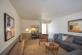 This single-floor residence offers a practical and comfortable layout across several well-defined living spaces. The living room serves as the heart of the home, featuring wall-to-wall carpeting, a textured ceiling, and a two-tone wall treatment with a neutral gray lower half. Natural light filters in through windows dressed with curtains, creating a warm and inviting atmosphere. The space is open enough to comfortably accommodate a sectional sofa, accent chairs, and a central coffee table alongside a media console.

The kitchen is efficiently designed with gray cabinetry, granite-style countertops, white appliances including a coil-top range and refrigerator, and a classic subway tile backsplash. Wood-look flooring runs throughout the kitchen, adding warmth and easy maintenance. Built-in shelving provides additional storage and display space.

The bedrooms are carpeted and generously sized, each featuring a sliding-door closet and windows with blinds that invite natural light. One bedroom includes a ceiling fan with a light fixture for added comfort. The bathroom is well-equipped with a vanity featuring granite-style counters, gray cabinetry, wood-look flooring, and a full tub and shower combination with gray tile surround and a privacy window above.