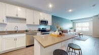 This single-floor residence offers a thoughtfully designed open-concept layout with seamless flow between the kitchen, dining, and living areas. The kitchen is a standout feature, showcasing crisp white shaker cabinetry, a classic subway tile backsplash, neutral-toned countertops, and a full suite of stainless steel appliances including a range, microwave, dishwasher, and refrigerator. A central island with seating provides additional workspace and casual dining options. Light wood-tone flooring runs throughout the main living areas, complementing the soft, neutral color palette accented by a calming teal wall.

The living area is bright and welcoming, benefiting from a large window that introduces natural light and outdoor views. Subtle sage-toned walls create a serene ambiance, making the space feel relaxed and comfortable.

The bedroom is generously sized with plush carpeting, elevated ceilings, and a large window that continues the theme of natural light. A spacious walk-in closet off the bedroom features built-in shelving and wire rack systems, offering ample organized storage.

The bathroom is finished with white cabinetry, a large framed mirror, and coordinating neutral countertops, maintaining the clean, cohesive aesthetic found throughout the residence.