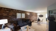 This single-floor space is defined by a prominent exposed brick wall that stretches across the living and dining areas, adding warmth, texture, and an industrial-chic character to the interior. The rich, dark-toned brickwork serves as a bold architectural backdrop, complementing the neutral palette of the surrounding walls and smooth white ceiling.

The living area is anchored by a substantial dark sectional sofa paired with a rectangular coffee table, creating a comfortable and functional seating arrangement. Framed artwork displayed against the brick wall adds a decorative touch, while a floor lamp provides supplemental lighting. The space flows seamlessly into the dining area, which features a round glass-top table surrounded by sleek chairs, illuminated by a chandelier overhead. A decorative wall-mounted piece accents the dining area's corner.

Plush, light-toned carpeting runs throughout, softening the contrast of the rugged brick and contributing to the overall cozy ambiance. The open layout between the living and dining zones promotes a sense of spaciousness, making this a well-connected and visually cohesive single-level space.
