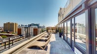 This expansive rooftop terrace offers a sophisticated urban retreat elevated above the surrounding cityscape. The deck is finished with light-toned wood-plank tiles that lend a clean, modern aesthetic throughout the space. A series of elegantly curved, sculptural lounge chairs are arranged along the terrace perimeter, each paired with a small round side table, creating a generous sunbathing area that takes full advantage of open sky exposure and panoramic city views.

A dedicated outdoor dining area is situated toward one end of the terrace, furnished with a long rectangular table and matching contemporary chairs, framed by slatted wood privacy screens and tall potted cypress trees that add a touch of greenery and definition to the space. Large planters with manicured trees are positioned throughout, providing natural accents against the neutral palette of the deck and building facade.

Along one side of the terrace, a continuous wall of floor-to-ceiling glass doors with dark frames provides direct access to the interior, filling the space with a sense of transparency and connection between indoors and out. A sleek metal railing runs along the open perimeter, ensuring unobstructed views of the surrounding urban skyline in multiple directions. The terrace receives abundant natural light throughout the day, making it an ideal setting for both relaxation and outdoor gatherings.