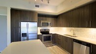 This single-floor space showcases a thoughtfully designed, modern interior with a strong emphasis on clean lines and contemporary finishes. The kitchen is a standout feature, offering a highly functional and stylish layout with full stainless steel appliances, including a French door refrigerator with water and ice dispenser, an electric range with over-the-range microwave, and a double-basin undermount sink. Rich, dark wood-toned cabinetry extends from floor to ceiling, providing generous storage while contributing to the space's sophisticated aesthetic. Light quartz countertops offer a striking contrast against the darker cabinetry, and a classic white subway tile backsplash adds a timeless yet modern touch. Recessed track lighting illuminates the kitchen workspace effectively, highlighting the quality of the finishes throughout. The light-toned hardwood flooring extends across the space, lending warmth and continuity to the overall design. The layout is efficient and well-organized, making excellent use of the available space. The combination of high-quality materials and modern design elements creates a cohesive, polished ambiance throughout this well-appointed residence.