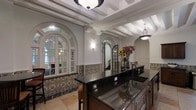 This single-story space showcases a distinctive Spanish Colonial architectural style, blending ornate detailing with functional design. The kitchen area serves as a focal point, featuring rich dark granite countertops set into wood cabinetry, complemented by an undermount sink. A decorative band of hand-painted, multi-colored encaustic tiles lines the lower walls, adding an authentic Mediterranean character to the space.

The ceiling draws immediate attention with its exposed white-painted beams arranged in a coffered pattern, enhancing the architectural depth of the room. Pendant light fixtures with dark metal accents hang from the ceiling, providing ambient illumination that complements wall-mounted sconces.

Arched French doors and arched passageways are recurring architectural elements throughout the space, flooding the interior with natural light while maintaining a sense of visual continuity. The flooring consists of large-format terra cotta-toned tiles, reinforcing the Mediterranean aesthetic. Textured plaster walls in a warm white finish contribute to the overall Old World ambiance, creating a cohesive and elegantly detailed interior environment.