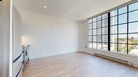 This single-floor unit offers an efficiently designed open-concept layout that combines the kitchen and living area into one cohesive space, complemented by a full bathroom. The living area is defined by its crisp white walls, light-toned hardwood flooring, and recessed lighting throughout. A standout feature is the large, multi-pane industrial-style window wall that spans nearly floor to ceiling, flooding the space with natural light and offering expansive urban skyline views. Exposed metallic ductwork runs along the upper portion of the space, lending an industrial aesthetic to the otherwise clean, minimalist design. A baseboard heating unit runs beneath the windows for added comfort. The kitchen is outfitted with medium-toned wood cabinetry accented by matte black hardware, a white subway tile backsplash, light stone countertops, and a full suite of stainless steel appliances, including a refrigerator, range, built-in microwave, and dishwasher. The bathroom features large-format neutral tile flooring, a wall-mounted toilet, and a generously sized glass-enclosed walk-in shower, maintaining the space's modern and clean aesthetic throughout.