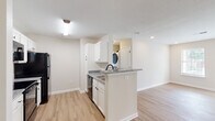 This single-floor residence offers a clean, modern aesthetic with a thoughtful layout designed for comfortable everyday living. The open-concept kitchen is a standout feature, showcasing white shaker-style cabinetry, granite countertops, a peninsula with a built-in sink, and black appliances including a refrigerator, range, dishwasher, and over-the-range microwave. Light wood-look flooring runs throughout the main living areas, enhancing the bright and airy feel of the space. The kitchen flows seamlessly into the living area, which benefits from recessed lighting and direct access to the exterior through a glass-paneled door flanked by windows that invite generous natural light. A dedicated in-unit laundry area is conveniently accessible from the kitchen. The bedroom is carpeted for comfort and features a ceiling fan with light fixture, a windowed wall that fills the room with sunlight, and a spacious reach-in closet. The bathroom is finished with consistent granite countertops, white cabinetry, a single-basin vanity with a large mirror, and matte black hardware and accessories. The overall design maintains a neutral, cohesive palette of whites and warm beiges, creating a fresh and inviting atmosphere throughout.