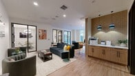 Located at 808 Chapel Street in the heart of New Haven, CT, this thoughtfully designed amenity floor offers a versatile and contemporary environment across seven distinct functional areas. The open lounge is anchored by plush navy blue sofas and accent chairs arranged over patterned area rugs atop warm hardwood-style flooring, creating a welcoming and social atmosphere. Recessed lighting throughout maintains a clean, bright ambiance. A dedicated business center features a built-in wood-paneled workstation with desktop computers, complemented by casual seating and direct access to an outdoor terrace. The terrace itself is a tranquil retreat, equipped with seating amid neatly arranged topiary planters against an exposed brick backdrop. Multiple private meeting and conference rooms provide focused collaboration spaces, each appointed with light wood tables, upholstered seating, and a signature plaid accent wall. Pendant lighting adds a stylish touch to each room. Individual focus pods enclosed in glass and dark steel framing offer quiet, single-occupancy workstations. A service nook with warm tile accents houses printing and copying equipment for added convenience. The overall design balances professional functionality with residential comfort throughout.
