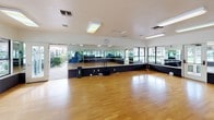 This aerobics room is a well-appointed, single-level fitness space designed to accommodate a variety of group exercise and movement activities. The room's most prominent feature is its expansive hardwood flooring, which provides a smooth, resilient surface ideal for aerobics, dance, yoga, and similar activities. Full-length mirrors line multiple walls, visually enlarging the space while allowing participants to monitor their form during workouts. A raised platform with a textured diamond-plate base runs along the mirrored walls, adding a functional and distinct design element to the room.

Large windows along one side of the room offer views of an outdoor pool area, allowing generous natural light to fill the space and creating an open, energizing atmosphere. Flush fluorescent ceiling fixtures provide consistent overhead lighting throughout. Multiple entry points, including glass-panel doors leading to the outdoors, enhance accessibility and airflow. A wall-mounted clock and available fitness equipment such as exercise mats are present to support instructor-led sessions. The overall design prioritizes functionality, visibility, and a bright, motivating environment suited to active fitness use.