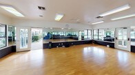 This aerobics room is a well-appointed, single-level fitness space designed to accommodate a variety of group exercise and movement activities. The room's most prominent feature is its expansive hardwood flooring, which provides a smooth, resilient surface ideal for aerobics, dance, yoga, and similar activities. Full-length mirrors line multiple walls, visually enlarging the space while allowing participants to monitor their form during workouts. A raised platform with a textured diamond-plate base runs along the mirrored walls, adding a functional and distinct design element to the room.

Large windows along one side of the room offer views of an outdoor pool area, allowing generous natural light to fill the space and creating an open, energizing atmosphere. Flush fluorescent ceiling fixtures provide consistent overhead lighting throughout. Multiple entry points, including glass-panel doors leading to the outdoors, enhance accessibility and airflow. A wall-mounted clock and available fitness equipment such as exercise mats are present to support instructor-led sessions. The overall design prioritizes functionality, visibility, and a bright, motivating environment suited to active fitness use.