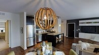 This single-floor space showcases a thoughtfully designed open-concept layout that seamlessly connects the kitchen and dining areas, creating a cohesive and inviting living environment. The kitchen is outfitted with dark, bold cabinetry that contrasts elegantly against granite countertops and stainless steel appliances, including a full-size refrigerator. A central island provides additional workspace and storage, enhancing both functionality and style. Track lighting runs along the ceiling, complementing the warm hardwood flooring that flows throughout the space. The dining area is anchored by a standout spherical wood-slat pendant light, which serves as a dramatic focal point and adds an artistic, contemporary touch to the room. A dining table set with place settings sits beneath this fixture, making the space feel both polished and welcoming. The open layout is framed by white trim and columns that add architectural definition, while the dark accent walls contribute depth and a modern edge. Natural light filters into the space, enhancing the warm tones of the wood flooring and cabinetry. Overall, the space combines modern design elements with warm, inviting details to create a functional yet visually compelling interior.