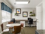 This business center is a thoughtfully designed, single-floor space comprising a well-appointed office and a connecting hallway. The office is efficiently organized to serve multiple purposes, featuring a long built-in workstation along one wall that accommodates multiple computer workstations with ergonomic task chairs. Opposite the work area, a round table with chairs provides a casual meeting or collaboration zone, while a pair of cushioned lounge chairs offers additional seating for informal gatherings.

The space is finished with neutral gray walls, crown molding, and a textured ceiling, lending it a polished, professional appearance. Patterned carpet in muted earth tones runs throughout, adding warmth underfoot. Natural light filters in through a window dressed with blue valance curtains and white blinds, brightening the interior and offering a view of the surrounding outdoor greenery.

Decorative wall-mounted shelves display small plants and décor items, while a curated selection of colorful framed artwork adds personality and visual interest to the walls. Overhead, recessed and flush-mounted lighting ensures consistent illumination throughout the day. The hallway connects the office to the broader building, providing easy access and a smooth flow through the space. Overall, the business center balances practicality with an inviting, creative atmosphere.