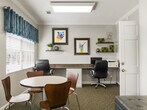 This business center is a thoughtfully designed, single-floor space comprising a well-appointed office and a connecting hallway. The office is efficiently organized to serve multiple purposes, featuring a long built-in workstation along one wall that accommodates multiple computer workstations with ergonomic task chairs. Opposite the work area, a round table with chairs provides a casual meeting or collaboration zone, while a pair of cushioned lounge chairs offers additional seating for informal gatherings.

The space is finished with neutral gray walls, crown molding, and a textured ceiling, lending it a polished, professional appearance. Patterned carpet in muted earth tones runs throughout, adding warmth underfoot. Natural light filters in through a window dressed with blue valance curtains and white blinds, brightening the interior and offering a view of the surrounding outdoor greenery.

Decorative wall-mounted shelves display small plants and décor items, while a curated selection of colorful framed artwork adds personality and visual interest to the walls. Overhead, recessed and flush-mounted lighting ensures consistent illumination throughout the day. The hallway connects the office to the broader building, providing easy access and a smooth flow through the space. Overall, the business center balances practicality with an inviting, creative atmosphere.