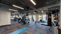 This fitness center combines industrial aesthetics with modern functionality across a generously sized single-floor layout. The exposed concrete ceiling and large cylindrical ductwork give the space a distinctive industrial character, while linear LED lighting ensures consistent illumination throughout. Colorful herringbone-patterned carpet tiles cover the floor, adding visual warmth that contrasts with the raw ceiling elements.

The facility is organized into distinct workout zones. The cardio area lines the perimeter windows, housing Echelon spin bikes and treadmills positioned to take full advantage of the tree-lined outdoor views through large, black-framed windows. A dedicated strength training section features cable machines, selectorized equipment, and weight benches, while a comprehensive dumbbell rack with a full weight range lines one wall alongside additional cable systems.

A stretching and functional training area provides yoga mats and stability balls of varying sizes, mounted neatly on storage racks. Wall-mounted televisions are positioned throughout the space for entertainment during workouts. The entry vestibule, framed by black-framed glass doors, provides a smooth transition into the main floor. Structural columns are integrated naturally into the open floor plan, maintaining a spacious and airy ambiance throughout.
