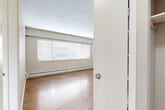 This one-bedroom, one-bathroom unit in New Westminster, BC offers a practical and well-organized single-floor layout. Warm hardwood flooring flows throughout the main living areas and bedroom, lending a classic character to the space. The bedroom is a comfortable size, featuring a wide horizontal window that admits natural light and offers views of the surrounding neighbourhood. A baseboard heating system runs along the walls, ensuring warmth throughout. The bedroom includes a mirrored sliding-door closet, providing convenient built-in storage. The kitchen is efficiently designed with medium-toned wood cabinetry, dark countertops, a tiled backsplash, and white appliances including a refrigerator and electric range. Light ceramic tile flooring grounds the kitchen area and connects seamlessly to the rest of the unit. The bathroom is finished with warm beige floor and wall tiles, a soaker tub with a tiled surround, a modern vessel sink set atop a dark-finished vanity, and a large mirror. A textured ceiling is present throughout the unit, adding a distinctive retro character. Overall, the space presents a clean, functional layout well-suited for comfortable everyday living.