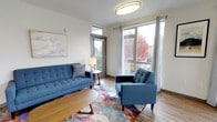 This single-floor living space offers a clean, contemporary design anchored by an abundance of natural light. The living area is defined by large floor-to-ceiling windows dressed with grommet-top curtains, framing lush outdoor views of mature trees displaying vivid autumn foliage. A glass-paneled door provides direct access to an outdoor balcony, further connecting the interior to the natural surroundings.

The flooring throughout is warm-toned hardwood, lending a grounded, inviting feel to the space. A flush-mount ceiling light provides consistent overhead illumination, complementing the generous daylight streaming in through the windows. The walls are finished in a crisp, neutral white, creating a bright and airy backdrop that enhances the sense of openness.

The living area is thoughtfully arranged with a tufted mid-century modern sofa and matching accent chair, both upholstered in a rich navy blue. A colorful abstract area rug adds a dynamic visual element, tying the seating arrangement together. A round coffee table and a small side table with a lamp complete the setup, contributing to a balanced and welcoming ambiance overall.