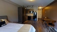 This single-level studio is designed for efficient, modern living within a compact and well-organized open-concept layout. The space seamlessly integrates the sleeping and kitchen areas, creating a cohesive and functional living environment. Dark hardwood flooring runs throughout, lending warmth and a contemporary aesthetic to the interior.

The kitchen is positioned along one wall and is fully equipped with dark wood cabinetry, a full-size refrigerator, a built-in microwave, a dishwasher, and a sink — providing all the essentials for everyday meal preparation. Track lighting overhead ensures the kitchen area is well-lit and practical.

The sleeping area occupies the adjacent open space, accommodating a full-size bed with easy access to the rest of the unit. A tall, dark wood wardrobe provides ample storage and serves as a visual anchor between the kitchen and living zones. Toward the entrance, a barn-style sliding door hints at additional storage or a closet space, adding a stylish architectural detail. The entryway is well-defined and leads naturally into the main living area. Overall, the unit offers a clean, modern ambiance with thoughtful use of every inch of available space.