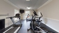 Located at 601 Northpointe Pky in Jackson, Mississippi, this fitness center is a well-appointed, single-floor facility designed to support a comprehensive workout routine. The space is finished with classic architectural details, including crown molding, wainscoting panels, and a textured ceiling, giving it a refined character uncommon in typical gym environments. Dark rubber flooring spans the entirety of the room, providing a durable, slip-resistant surface suited for exercise. Natural light flows in through multiple windows, offering views of the surrounding outdoor greenery and keeping the space bright and inviting. A wall-mounted flat-screen television is positioned for easy viewing from the cardio area, enhancing the workout experience. The cardio section is equipped with multiple treadmills and an elliptical trainer, while a dedicated strength area houses Cybex weight machines, including a functional trainer and additional resistance equipment. A classic brick fireplace with a white mantle adds a distinctive architectural focal point. French-style glass doors connect to an adjacent area, contributing to an open, airy feel. A sanitizing station is also present, reflecting attention to cleanliness and member convenience. Overall, this fitness center balances practical functionality with a polished, welcoming aesthetic.