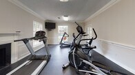 Located at 601 Northpointe Pky in Jackson, Mississippi, this fitness center is a well-appointed, single-floor facility designed to support a comprehensive workout routine. The space is finished with classic architectural details, including crown molding, wainscoting panels, and a textured ceiling, giving it a refined character uncommon in typical gym environments. Dark rubber flooring spans the entirety of the room, providing a durable, slip-resistant surface suited for exercise. Natural light flows in through multiple windows, offering views of the surrounding outdoor greenery and keeping the space bright and inviting. A wall-mounted flat-screen television is positioned for easy viewing from the cardio area, enhancing the workout experience. The cardio section is equipped with multiple treadmills and an elliptical trainer, while a dedicated strength area houses Cybex weight machines, including a functional trainer and additional resistance equipment. A classic brick fireplace with a white mantle adds a distinctive architectural focal point. French-style glass doors connect to an adjacent area, contributing to an open, airy feel. A sanitizing station is also present, reflecting attention to cleanliness and member convenience. Overall, this fitness center balances practical functionality with a polished, welcoming aesthetic.