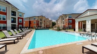 This outdoor amenity space centers around a generously sized resort-style swimming pool with clear blue water, surrounded by a spacious concrete deck finished in warm neutral tones. The pool area is well-equipped with rows of black chaise lounge chairs positioned along both sides, offering ample room for sunbathing and relaxation. Umbrella-shaded seating areas are also visible toward the rear of the deck, providing shaded options for residents seeking respite from the sun. The pool itself features clean tile borders and appears well-maintained, reflecting the surrounding sky. Enclosing the space is a black metal perimeter fence that defines the amenity area while maintaining an open, airy feel. The surrounding multi-story residential buildings, clad in a combination of deep red and neutral tan exterior finishes with dark rooflines, frame the pool courtyard on multiple sides, creating a sense of enclosure and community. Mature trees are visible in the background, softening the built environment and adding natural greenery. The overall setting conveys a vibrant, community-focused outdoor living experience designed for leisure and social activity.